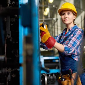 Confident young engineer wearing overall and checked shirt looking away while carrying out inspection at manufacturing plant, blurred background